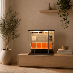 May include: A miniature model of a subway car interior featuring four bright orange seats, vertical silver support poles, a rectangular window, and interior lighting. The model includes small wall advertisements and a black base, replicating a train carriage.