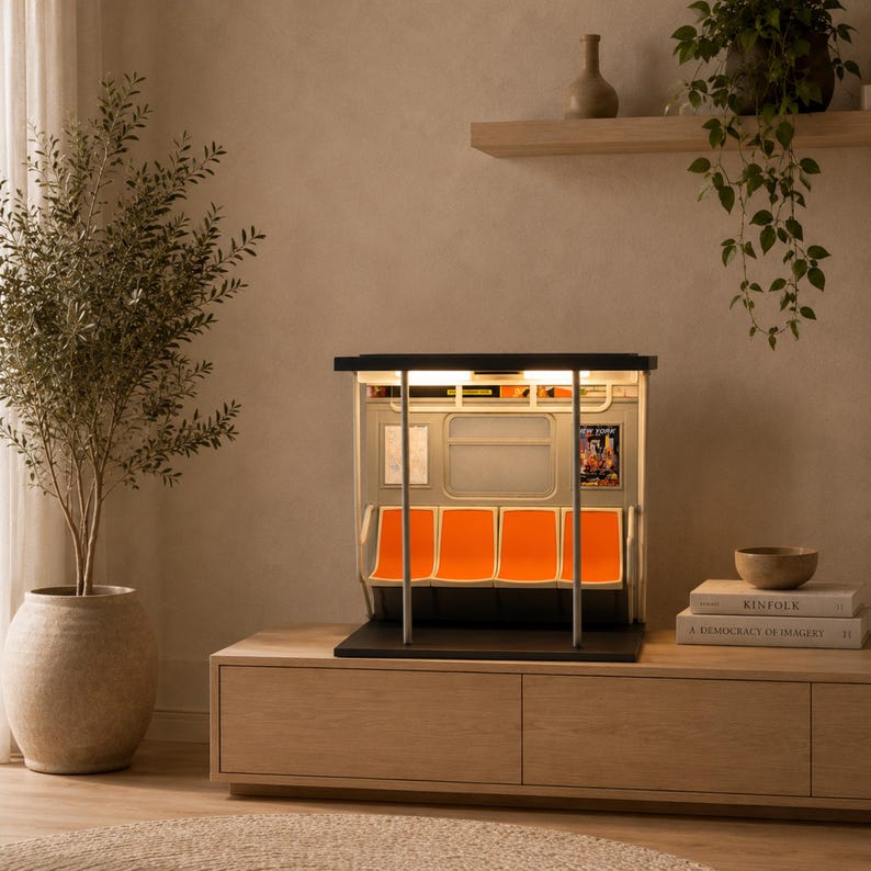 May include: A miniature model of a subway car interior featuring four bright orange seats, vertical silver support poles, a rectangular window, and interior lighting. The model includes small wall advertisements and a black base, replicating a train carriage.