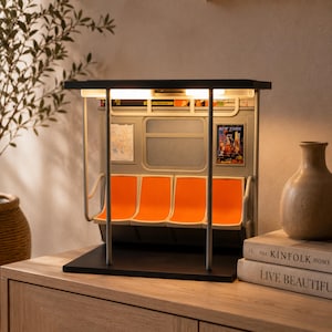 May include: A miniature subway car interior lamp featuring four bright orange molded seats, vertical silver support poles, and a black base. The interior walls are light gray with a small New York poster and a map graphic, illuminated by overhead lights.
