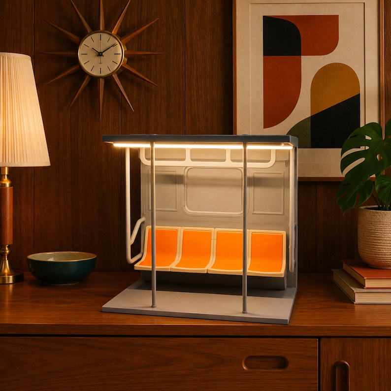 May include: A miniature subway car interior lamp featuring four bright orange molded plastic seats, a gray frame, and a white overhead grab rail. The structure is illuminated by a horizontal LED light strip integrated into the ceiling of the unit.