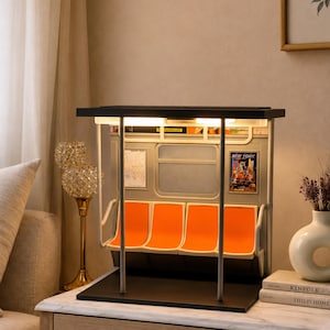 May include: A miniature model of a subway car interior featuring four bright orange molded seats, silver vertical support poles, a rectangular window, and a New York themed advertisement poster on the wall, all illuminated by an overhead light.