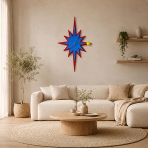 May include: A starburst-shaped wall clock with a blue centre and red points. The clock has a yellow sphere as a second hand. The clock is mounted on a neutral-coloured wall above a white sofa and round wooden coffee table.