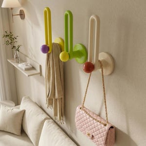 Pode incluir: Três cabides de parede coloridos em amarelo, verde e branco sujo, cada um com um design arredondado e uma bola contrastante na extremidade. Um cachecol bege e uma bolsa rosa acolchoada com uma corrente dourada estão pendurados nos cabides. Os cabides estão montados numa parede clara.