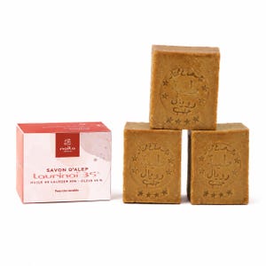 Peut inclure: Trois blocs rectangulaires de savon d'Alep brun avec du texte arabe et des étoiles estampés, accompagnés d'une boîte en carton rouge et blanc étiquetée Savon d'Alep Laurinaï 35% Huile de Laurier 35% - Olive 65% Peau très sensible.