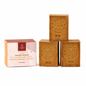 Peut inclure: Trois blocs rectangulaires de savon d'Alep brun empilés en pyramide, chacun estampillé de texte arabe et d'étoiles. À côté se trouve une boîte en carton carrée étiquetée Naila Nature Savon d'Alep Laurinaï 20% Huile de Laurier 20% - Olive 80%.