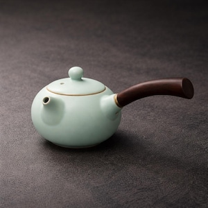 May include: A light green ceramic teapot with a dark brown wooden handle and lid. The teapot has a small spout and a round, bulbous shape. The lid has a small knob on top. The handle is curved and attached to the side of the teapot.