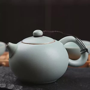 May include: A pale green ceramic teapot with a rounded body, curved handle, and a small spout. The lid has a small knob and a brown cord is attached to the handle. The teapot is sitting on a dark surface.