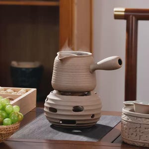 May include: A beige ceramic teapot with a side handle rests on a matching circular stove base featuring multiple cutouts. The textured, matte-finish teapot has a spout and emits visible steam, while the base is designed for heating.