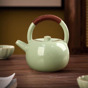 May include: A celadon green ceramic teapot with a rounded body and a curved handle wrapped in brown woven material. The teapot has a small spout and a lid with a small knob. A small teacup is visible in the background.