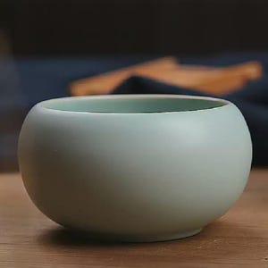 May include: A small, round, pale green ceramic bowl with a smooth, matte finish. The bowl has a slightly wider opening and a rounded base. The interior rim is a lighter shade of green. The bowl is sitting on a wooden surface.