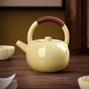 May include: A pale yellow ceramic teapot with a brown woven handle. The teapot has a rounded body, a small spout, and a lid with a small knob. The teapot is sitting on a wooden surface.