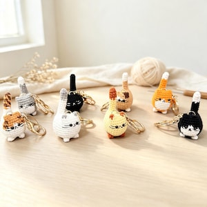 May include: A collection of eight small, round, crocheted cat keychains in various colours including black, white, orange, grey, and calico. Each cat features a tall, upright tail, embroidered facial details, and a gold-toned metal keyring.