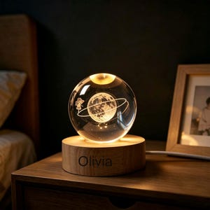 May include: A clear glass sphere lamp featuring an internal 3D laser-etched moon with a ringed planet and a small figure, resting on a circular light-colored wooden base with the name Olivia engraved in dark letters on the front side.