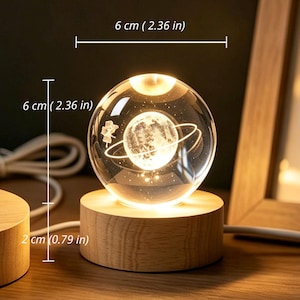 May include: A clear glass sphere night light featuring an internal 3D laser-etched planet with rings and a small figure, resting on a cylindrical light-colored wood base. The sphere measures 2.36 inches in diameter and the base is 0.79 inches tall.