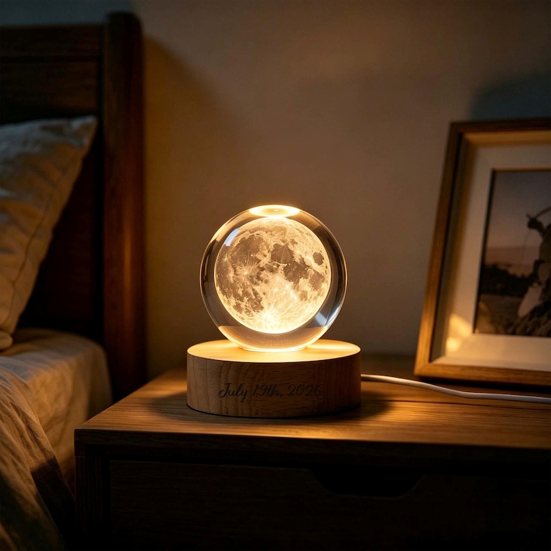 May include: A spherical glass lamp featuring a detailed 3D laser-engraved moon inside, resting on a circular light-colored wooden base. The base is engraved with the text July 19th, 2020. The lamp emits a warm, soft glow, illuminating the moon.
