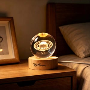 May include: A clear glass sphere containing a laser-etched 3D model of the planet Saturn with its rings, mounted on a circular light-colored wooden base. The base features the name William engraved in a dark script font on the front surface.
