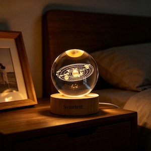 May include: A clear glass sphere lamp featuring an internal laser-etched solar system design. The sphere rests on a cylindrical light-colored wooden base with the name Scarlett engraved on the front, emitting a warm glow from within.