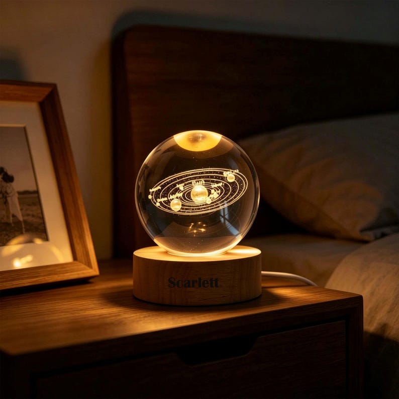 May include: A clear glass sphere lamp featuring an internal laser-etched solar system design. The sphere rests on a cylindrical light-colored wooden base with the name Scarlett engraved on the front, emitting a warm glow from within.