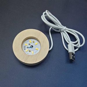 May include: A circular light-colored wooden display base featuring a central recessed circular LED light component with six small yellow diodes and a white circuit board. A white power cord with a USB-A connector is attached to the side.