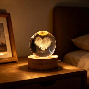 May include: A clear glass sphere containing a glowing, textured heart shape, mounted on a cylindrical wooden base. The base features the engraved text "Forever starts with you." The sphere emits a warm, soft light, illuminating the heart design.
