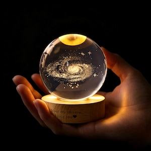 May include: A clear glass sphere containing a laser-etched spiral galaxy and stars, illuminated from below by a round wooden base. The base features the engraved text "Your my universe, I love you" followed by a small heart symbol in black ink.