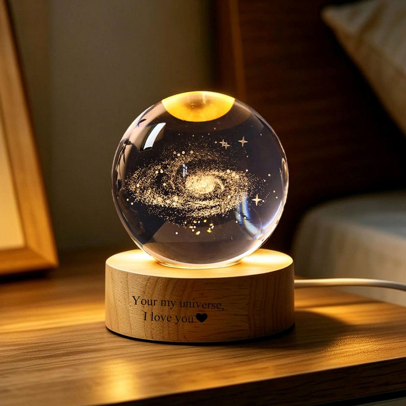 May include: A clear glass sphere featuring an internal laser-etched spiral galaxy design, resting on a cylindrical light-colored wooden base. The base is engraved with the text "Your my universe, I love you" followed by a small heart symbol.
