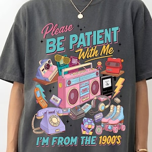 Puede incluir: Camiseta gris oscuro con un gráfico colorido que presenta elementos retro como una radio, patines y un teléfono de disco. El texto dice "Please Be Patient With Me I'm From The 1900's."