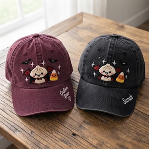 May include: Two baseball caps, one maroon and one black, featuring embroidered designs of a smiling dumpling character with bat wings, candy corn, bats, and stars. The maroon cap has the text Custom Boo and a heart, while the black cap says Sarah.