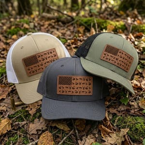 May include: Three baseball caps in tan, charcoal gray, and olive green, each featuring a rectangular leather patch on the front. The patches display a flag design composed of small silhouettes of deer and wildlife, with mesh back panels.