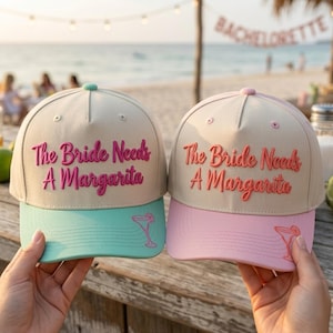 May include: Two beige baseball caps with embroidered text reading The Bride Needs A Margarita. One cap features a mint green brim with a small margarita glass icon, while the other has a light pink brim with a matching margarita glass icon.