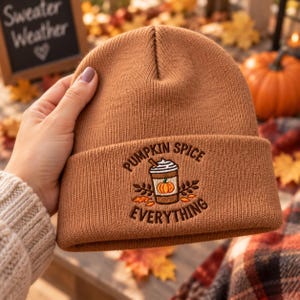May include: A solid brown knit beanie featuring an embroidered design on the cuff. The embroidery depicts a coffee cup with whipped cream and a small pumpkin, surrounded by autumn leaves, with the text PUMPKIN SPICE EVERYTHING arched around it.