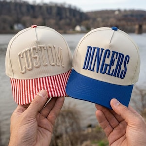 May include: Two five-panel baseball caps with off-white crowns. The left cap features the word CUSTOM in raised off-white embroidery and a red and white vertical striped brim. The right cap features the word DINGERS in blue embroidery and a blue brim.