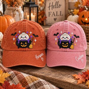 May include: Two baseball caps in orange and pink, each featuring an embroidered graphic of a smiling dumpling inside a black jack-o'-lantern cauldron with purple liquid, bats, and a broom. The orange cap says Harper, the pink cap says Custom.