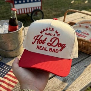 May include: A white and red baseball cap with the text "MAKES ME WANT A Hot Dog REAL BAD" embroidered in red. The cap has a red brim and a red button on top. The image suggests a summer picnic or outdoor event.