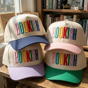 May include: Four baseball caps with the word "BOOKISH" embroidered in rainbow colors. The caps have different colored brims: navy blue, pink, green, and lavender. The caps are beige.