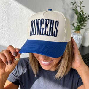 May include: A cream-colored baseball cap with a blue brim and the word "DINGERS" embroidered in navy blue. The cap has a structured fit and a button on top. The hat is being held by a person.