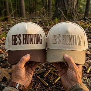 May include: Two baseball caps with a structured crown and a curved brim. The left cap is cream with a dark brown brim and dark brown embroidered text reading HE'S HUNTING. The right cap is cream with a tan brim and tan embroidered text reading HE'S HUNTING.