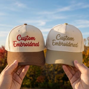 May include: Two baseball caps with off-white crowns. The left cap features a brown brim and red embroidered text reading Custom Embroidered. The right cap features a tan brim and gold embroidered text reading Custom Embroidered in cursive script.
