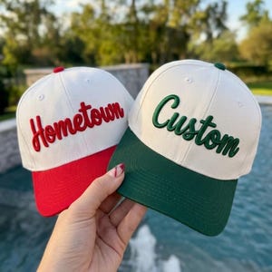 May include: Two baseball caps with white crowns. The left cap features red embroidery reading "Hometown" and a red brim. The right cap features dark green embroidery reading "Custom" and a dark green brim. Both caps have a button on top.