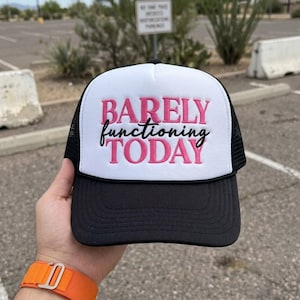 May include: A trucker hat with a white foam front panel, black mesh back panels, and a black brim. The front features embroidered text in pink and black that reads BARELY functioning TODAY, with the word BARELY and TODAY in pink block letters.