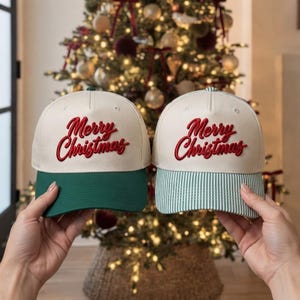 May include: Two baseball caps with off-white crowns and red embroidered text reading Merry Christmas. The left cap features a solid dark green brim, while the right cap has a light green and white pinstriped brim. Both have structured panels.