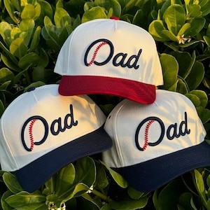 May include: Three white baseball caps with the word "Dad" embroidered in navy blue script, with a baseball design. One cap has a red brim, and the other two have navy blue brims. The caps are set against a background of green foliage.