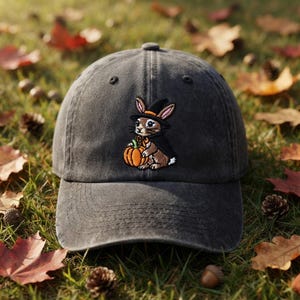 May include: A charcoal gray baseball cap featuring an embroidered patch of a brown rabbit wearing a black witch hat and cape while holding a small orange pumpkin. The cap has a distressed, washed-out fabric texture and a curved brim.