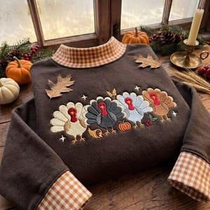 May include: A dark brown long-sleeve sweatshirt featuring a mock neck and sleeve cuffs in an orange and white gingham pattern. The chest displays an embroidered design of four turkeys in different colors, a small pumpkin, and wheat.