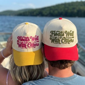 May include: Two off-white baseball caps with red buttons on top. The left cap has a yellow brim and pink embroidered text reading Floats Well With Others. The right cap has a red brim and green embroidered text reading Floats Well With Others.