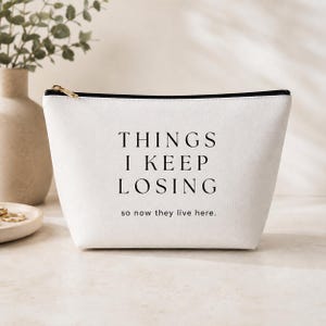 May include: A white zippered pouch featuring the black text THINGS I KEEP LOSING and the smaller text so now they live here. The bag has a black zipper closure along the top edge and a clean, structured design for storing various small items.