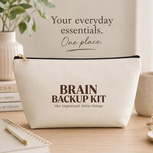 May include: A cream-colored canvas zippered pouch featuring the dark brown text BRAIN BACKUP KIT and the important little things printed on the front. The pouch has a black zipper closure and a gold-toned metal zipper pull.