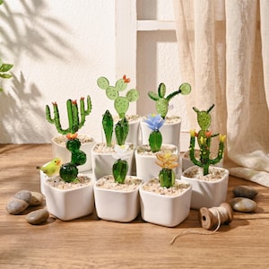 Peut inclure: Collection de sculptures de cactus en verre dans de petits pots carrés blancs. Les cactus sont de différentes nuances de vert, certains avec de petites fleurs colorées. Une figurine de petit oiseau jaune est posée près d'un pot.