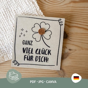 Könnte beinhalten: Eine quadratische, hellbraune Karte mit einem vierblättrigen Kleeblatt-Design und dem deutschen Text "GANZ VIEL GLÜCK FÜR DICH!". Eine Münze befindet sich in der Mitte des Kleeblatts. Die Karte liegt auf einer Holzoberfläche.