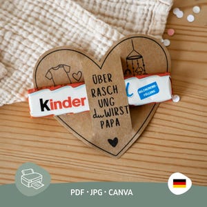 Puede incluir: Una tarjeta en forma de corazón de papel marrón con el texto alemán "Überraschung, du wirst Papa" y una barra de chocolate Kinder. La tarjeta tiene un gráfico de ropa de bebé y una ilustración de móvil. También se ven las palabras "PDF, JPG, CANVA".